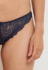 Perizoma in pizzo blu navy con motivi floreali intricati, bordi ondulati e una morbida fascia in vita, che esalta un design delicato.