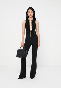 Woman in black sleeveless top with gold buttons, black corset belt, black pants, holding a black handbag with logo.