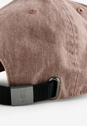 Ruskea corduroy-hattu, jossa on kaareva lippa, musta säädettävä hihna ja metallinen yksityiskohta, jossa on vuohen kallon kuvasto.
