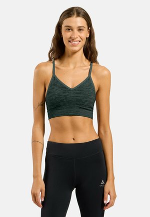 Femme aux longs cheveux souriante, portant un soutien-gorge de sport vert foncé et un legging taille haute noir, sur un fond clair uni.