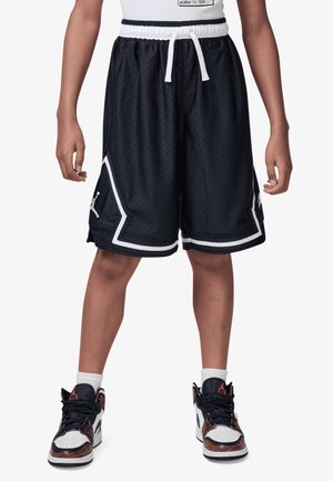 Osoba ubrana w czarne szorty do koszykówki z białym wykończeniem oraz czarno-biało-czerwone wysokie buty sportowe, stojąca z rozstawionymi nogami.