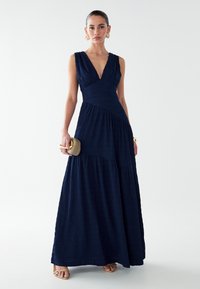 Marineblaues Maxikleid mit tiefem V-Ausschnitt, strukturiertem Stoff, figurbetontem Oberteil und ausgestelltem, gestuftem Rock, kombiniert mit goldenen Accessoires.