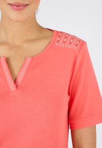 Koraalkleurig katoenen T-shirt met een V-hals afgewerkt met decoratieve stiksels en textuuraccenten op de schouders. Korte mouwen.