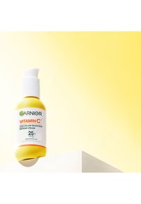 Gul flaske med Garnier Vitamin C 2-i-1 glow booster serumcreme med SPF 25 på en hvid overflade mod en gradient gul baggrund.