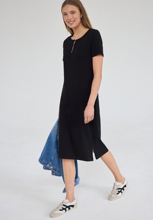 Jeune femme souriante, marchant, portant une robe midi noire à manches courtes et des baskets blanches, tenant une veste en jean bleue dans une main.