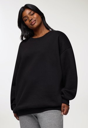 Donna con lunghi capelli neri, che indossa un ampio sudario nero e pantaloni grigi, in piedi contro uno sfondo chiaro e semplice.