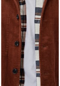 Bruine corduroy jas over een wit overhemd, gecombineerd met een wit, bruin en marineblauw geruit overhemd met knopen.