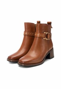 Botas de tobillo de cuero marrón con punta cuadrada, cierre de cremallera, correa decorativa con tachuelas doradas y tacón cuadrado. Textura suave.