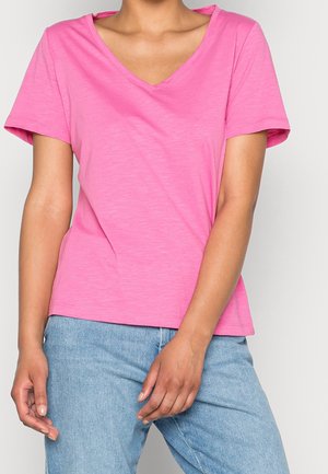 Osoba nosząca jaskraworóżowy T-shirt z krótkim rękawem i dekoltem w serek oraz jasnoniebieskie jeansy, stoi z rozluźnionymi rękami.