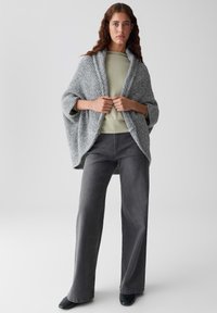 Frau in einer grauen Strickjacke über einem hellgrünen Oberteil, dunkelgrauen weitem Jeans und schwarzen Schuhen, die vor einem einfarbigen Hintergrund steht.