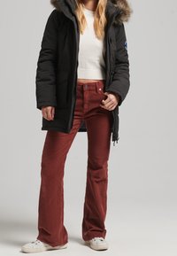 Black puffer jacket with fur-trimmed hood, paired with fitted corduroy pants in rust color and white sneakers. Zigzag texture visible on pants.