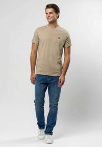 Beežist puuvillane T-särk ümarate kaelustega, lühikeste varrukatega ja väikese musta logoga vasakul rinnal, kombineeritud siniste teksade ja valgete tossudega.