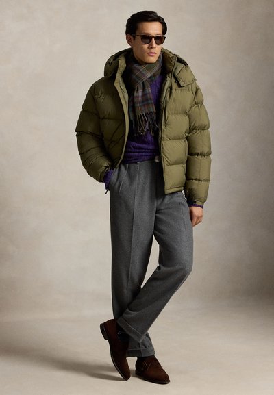 Homme portant des lunettes de soleil foncées, une doudoune vert olive, une écharpe à carreaux, un pull violet, un pantalon gris et des chaussures marron, debout avec une main dans la poche.