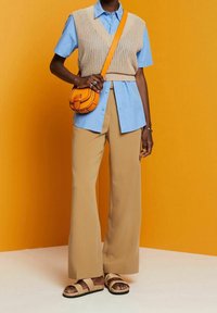Une personne portant un gilet en tricot beige par-dessus une chemise bleue, un pantalon large beige, des sandales beiges et portant un sac bandoulière orange devant un fond orange.
