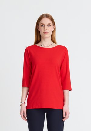 Frau mit langem, glattem Haar, die ein rotes Dreiviertelärmel-Top, dunkle Hosen, eine silberne Halskette und ein Armband trägt, steht vor einem schlichten weißen Hintergrund.