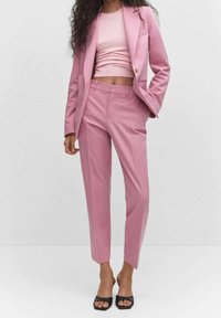 Femme portant un blazer rose ajusté et un pantalon assorti, avec un haut court rose et des talons ouverts noirs, posant devant un fond uni.