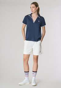 Polo shirt navy con bordi bianchi, pantaloni corti bianchi e calzini a righe multicolori. L'outfit presenta un design sportivo, con un piccolo logo sulla maglietta.