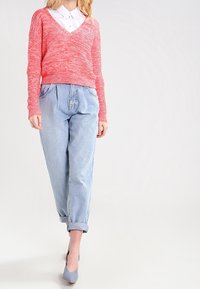 Maglione corallo lavorato a maglia con scollo a V, abbinato a pantaloni di denim azzurri con orli risvoltati. La modella indossa tacchi grigi, mostrando un outfit casual.