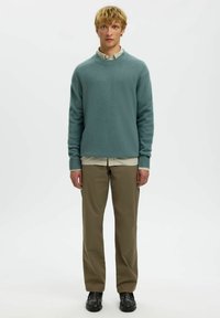 Pull léger teal avec encolure côtelée sur une chemise blanche à rayures, associé à un pantalon khaki et des chaussures noires, offrant une coupe décontractée.