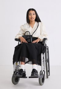 Utente di sedia a rotelle con una blusa crema e una gonna plissettata nera, che tiene una borsa nera testurizzata. Scarpe nere e calze bianche visibili.