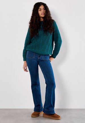 Maglione lavorato a maglia in teal con motivo a diamante testurizzato, abbinato a jeans svasati blu e pantofole in suede marroni. Un ensemble semplice e casual.
