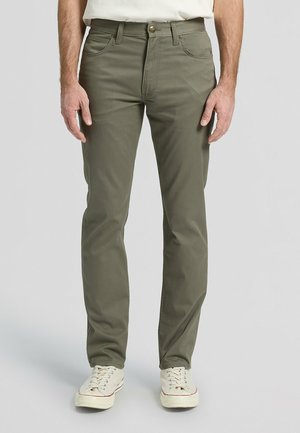 Mężczyzna w oliwkowozielonych spodniach o prostych nogawkach i białych trampkach z płótna, stojący na tle jasnoszarego tła.