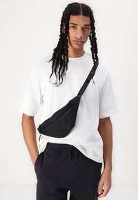 Black sling bag with a textured finish, featuring a zip closure and a strap labeled "JACK & JONES". Worn over a white t-shirt.