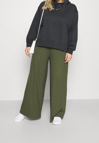 Mörkgrå huvtröja med dragsko, tillsammans med löst sittande olivgröna ribbade byxor. Vita sneakers och en liten vit väska kompletterar outfiten.