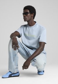Lys blå T-shirt med korte ærmer og et tekstureret mønster samt et grønt logo, parret med lys blå jeans og blå-sorte sneakers. Mørke solbriller.