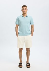 Polo shirt tricoté bleu ciel avec un motif texturé, manches courtes, associé à un short en lin crème et des sandales noires.