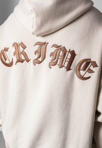 Cremefarbener Hoodie mit großem, kunstvollem braunen Stickmotiv, das "CRIME" auf dem Rücken spellt. Hergestellt aus weichem Stoff mit glatter Textur.