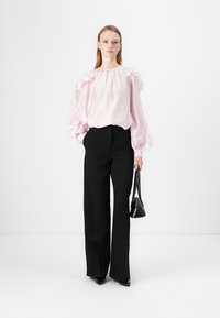 Blusa rosa chiaro con maniche a volant, abbinata a pantaloni neri a vita alta. La modella tiene una piccola borsa nera a tracolla. Tessuto morbido e vestibilità strutturata.