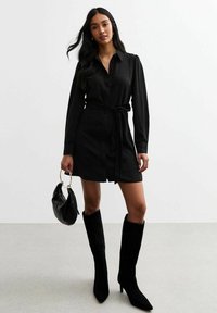 Abito nero con maniche lunghe, chiusura con bottoni davanti e cintura in vita. Completato da stivali neri in suede al ginocchio e una borsa nera.