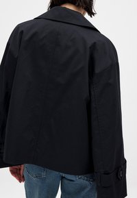 Svart kort jacka med breda slag och knäppta manschetter, tillverkad av ett slätt material. Jeans synliga nederst, enkel bakgrund.