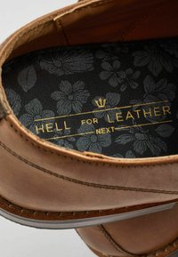 Unutrašnjost smešnih kožnih cipela s utisnutim cvjetnim uzorkom u crnoj boji. Zlatni tekst kaže "HELL FOR LEATHER NEXT." Vidljivo kremasto šivanje.