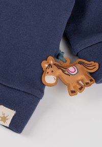 Marineblaues Baumwollkleidungsstück mit strukturiertem Finish, ausgestattet mit einem Cartoon-Pferde-Patch in Beige, das detaillierte Akzente in Rot, Grün und Schwarz aufweist.