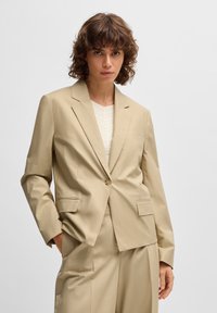 Beige räätälöity bleiseri, jossa on yksittäinen nappi, kaksi etutaskua ja sileä pinta. Yhdistetty tekstuuriseen kermaiseen paitoihin.