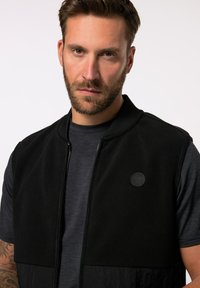 Schwarze ärmellose Jacke mit glatter Textur, Frontreißverschluss und einem runden Logo. Wird über einem strukturierten dunkelgrauen Hemd getragen.