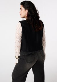 Zwarte, geribbelde top over een wit, kanten shirt met lange mouwen. De outfit wordt gecombineerd met donkergrijze, hooggesloten jeans.