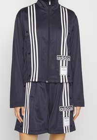 Marinblå idrottsbyxor med vita ränder, bestående av en zip-up jacka, tryckknappar och två logopatchar på shortsen.