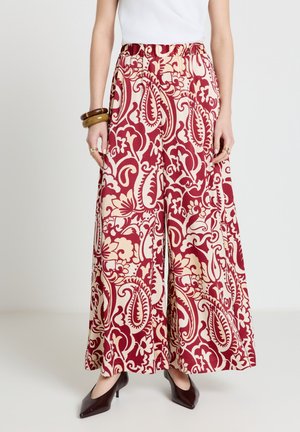 Donna che indossa pantaloni a gamba larga con motivo floreale paisley rosso e crema, abbinati a tacchi scuri a punta e braccialetti spessi.