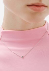 Collar de cadena de plata con un pequeño colgante redondo que tiene una piedra clara, llevado sobre una blusa ajustada de color rosa con un escote alto.
