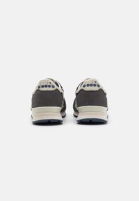 Graue Wildleder-Sneaker mit beigen Akzenten, die ein minimalistisches Design und blaue Gummisohlen aufweisen. Die Ferse verfügt über ein Markenlogo in kontrastierender Farbe.