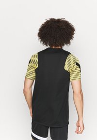 Svart sportströja med korta ärmar som har ett texturerat gult geometriskt mönster. Slätt tyg med rund halsringning och lös passform.