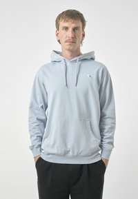 Light blue hoodie made of cotton with a front pocket, drawstring hood, and small bird logo on the left chest. Paired with black pants.
