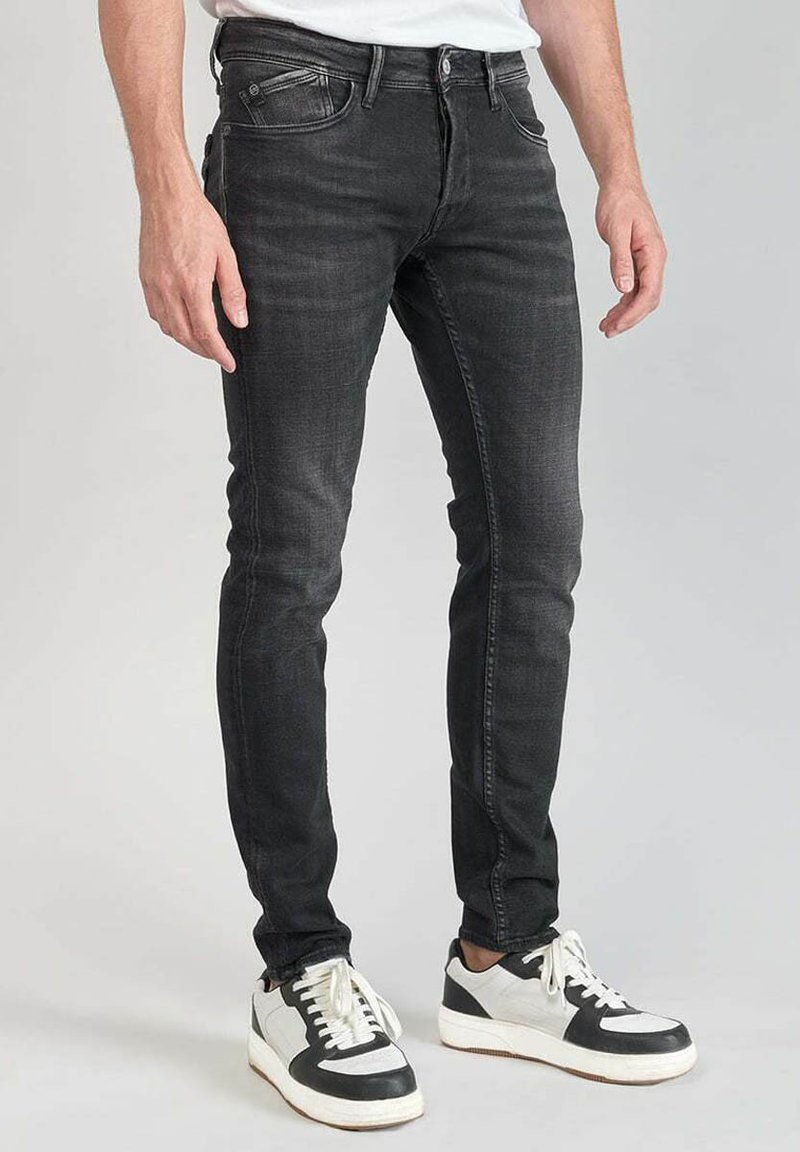 Jeans de corte ajustado en gris oscuro con una textura de denim suave, que cuentan con dos bolsillos frontales y un cierre estándar de botón y cremallera, combinados con zapatillas negras y blancas.