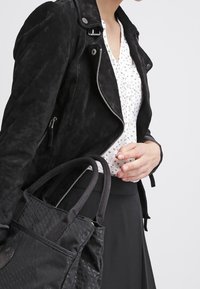 Musta samettinen moottoripyöräntakki, jossa on hopeiset yksityiskohdat, käytetty valkoisen kirjavan paidan päällä, yhdistetty mustaan teksturoituun käsilaukkuun.