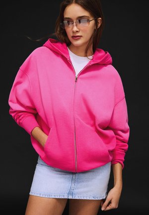 Femme portant des lunettes de soleil carrées translucides, un sweat à capuche zippé rose vif et une mini-jupe en jean clair sur un fond sombre.