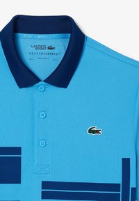 Blå pikétröja med marinblå krage, tre knappar och Lacoste krokodil-logotyp. Har horisontella ränder i marinblått på bröstet. Normal passform.