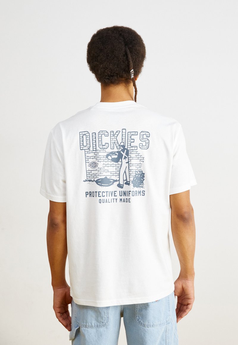 Balta medvilninė marškinė su mėlynu piešiniu ant nugaros, ant kurio yra tekstas "DICKIES PROTECTIVE UNIFORMS QUALITY MADE" ir figūra.